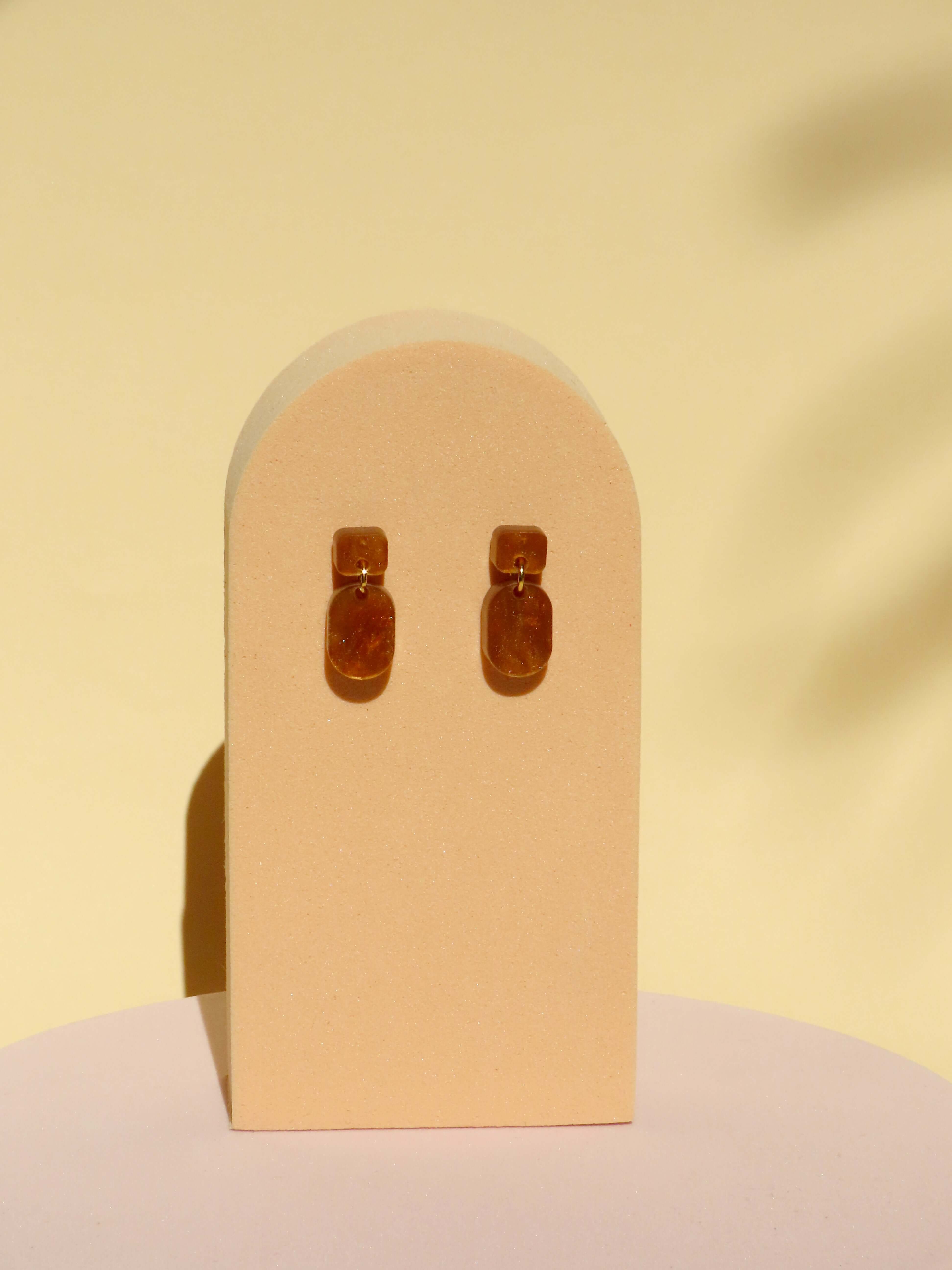 Paar kleiner Ohrhänger aus karamellbraunem, marmoriertem Acryl mit eckigem Stecker und ovalem Anhänger, verbunden durch ein goldenes Element
