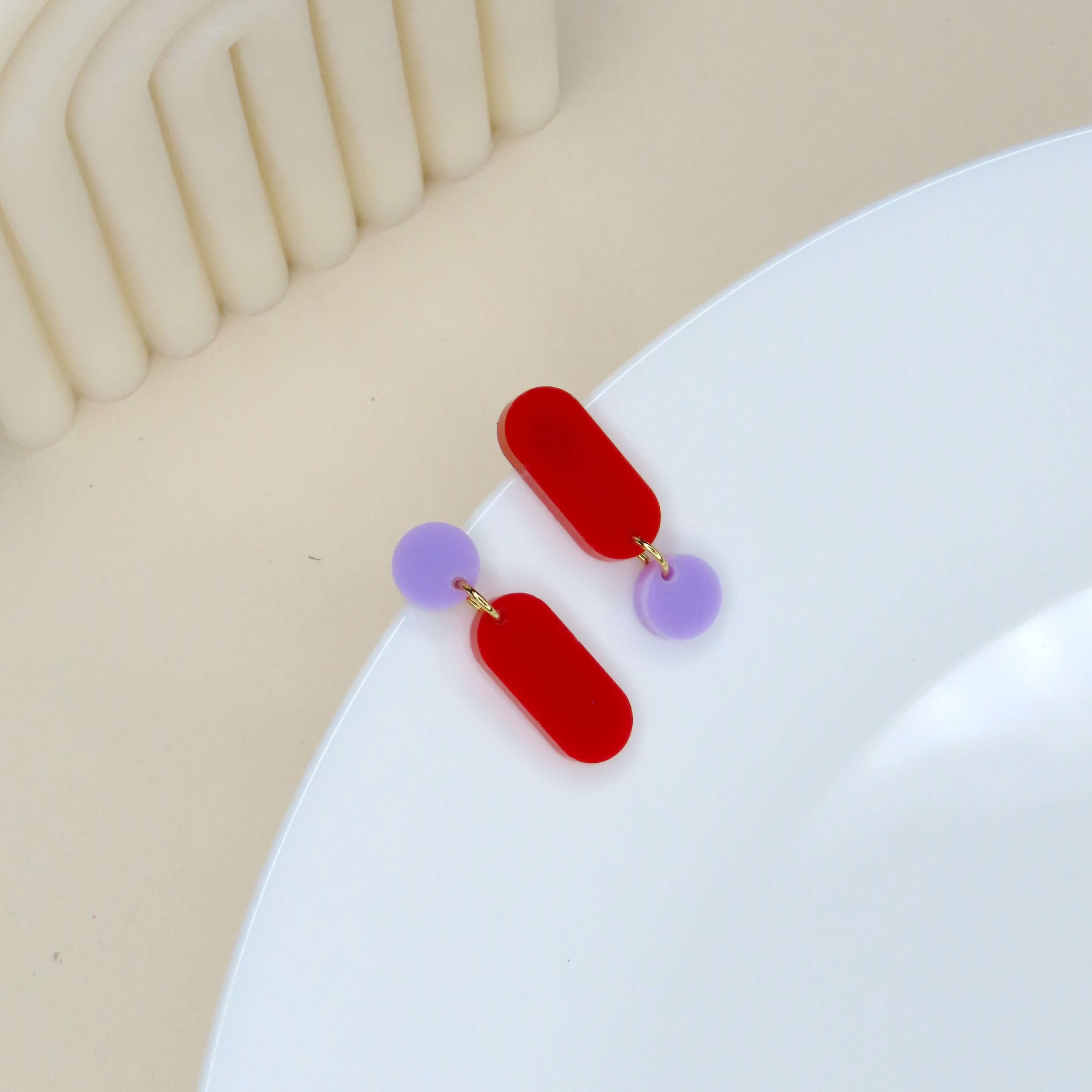 Asymmetrische Acryl Ohrringe in Flieder und Rot mit rundem und länglichem Element, verbunden durch kleine goldfarbene Ringe