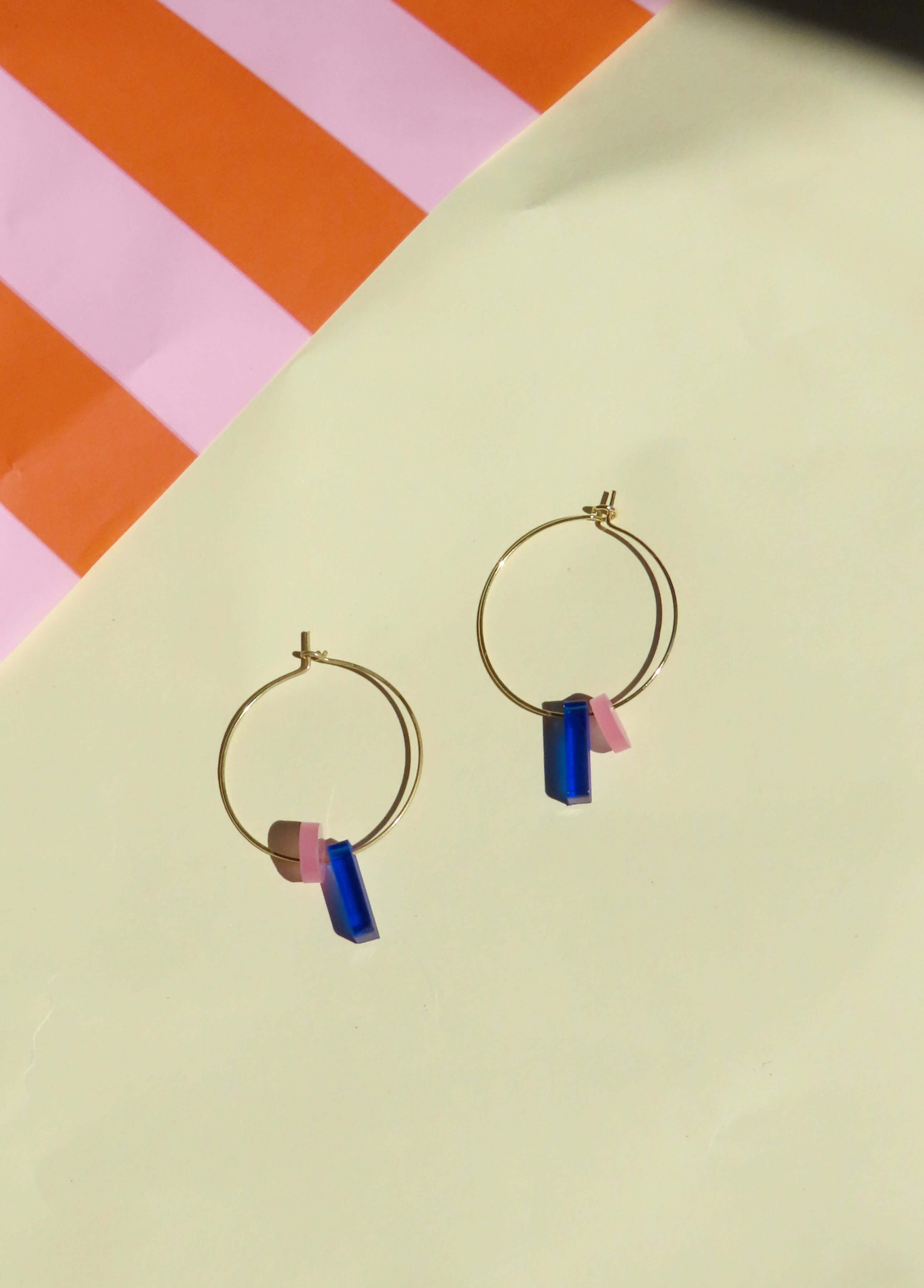 goldene creolen mit geometrischen anhängern in blau und rosa auf pastellfarbenem hintergrund