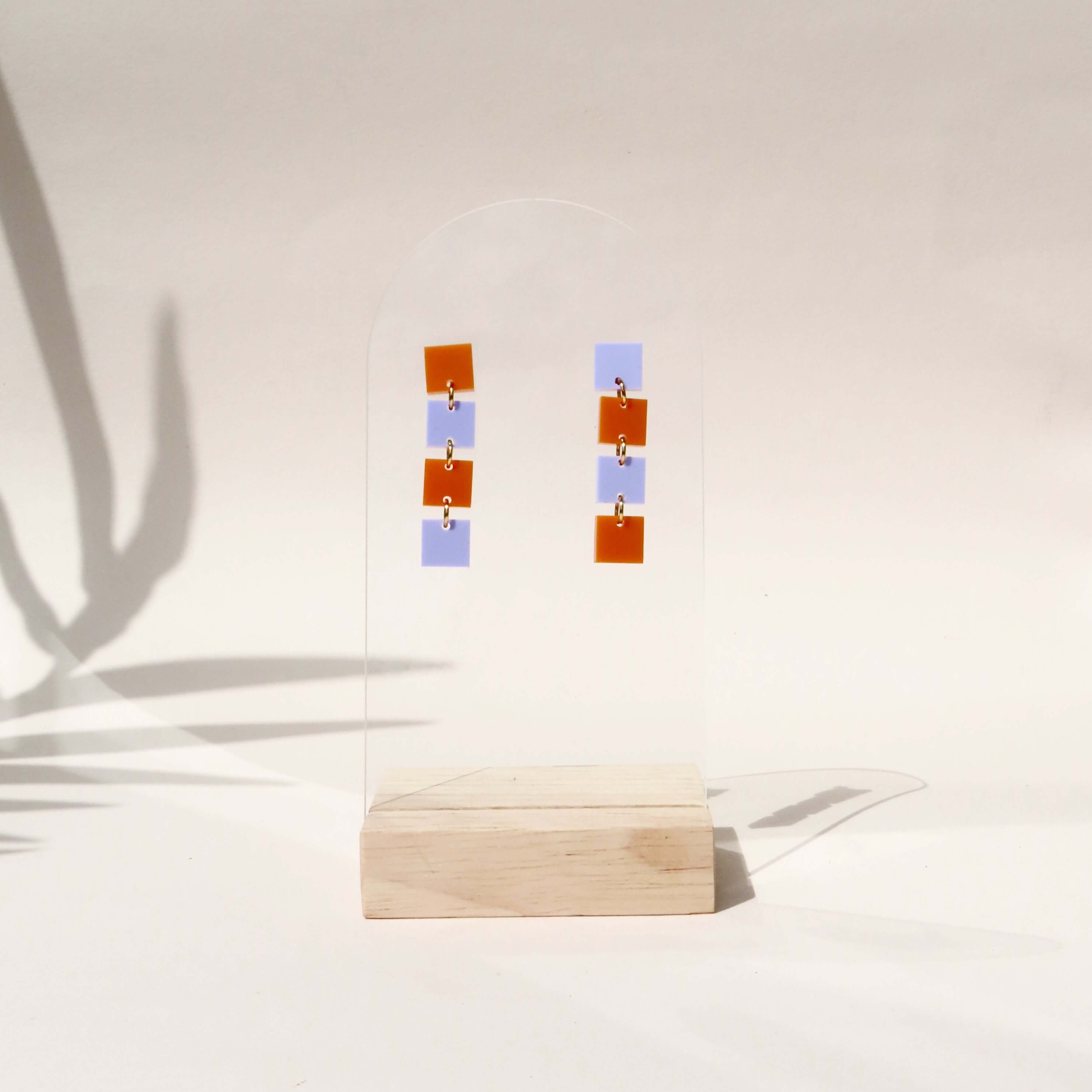 Hängende Statement-Ohrringe aus Acryl mit kleinen Quadraten in Orange und Flieder, verbunden durch goldfarbene Ringe.