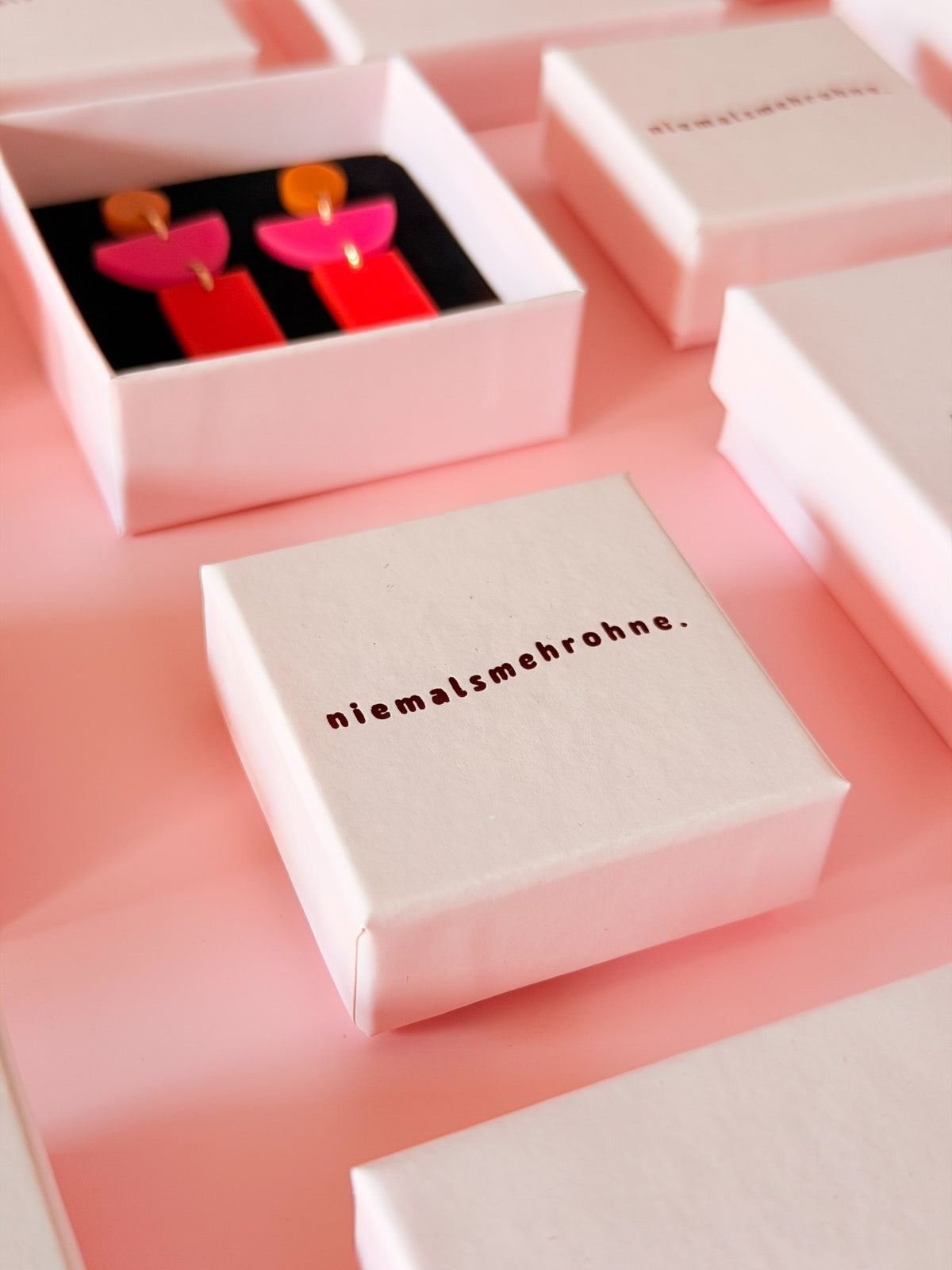 Weiße Schmuckschachtel mit niemalsmehrohne Logo auf rosa Hintergrund, umgeben von weiteren Geschenkboxen und farbenfrohen Acryl-Ohrringen.