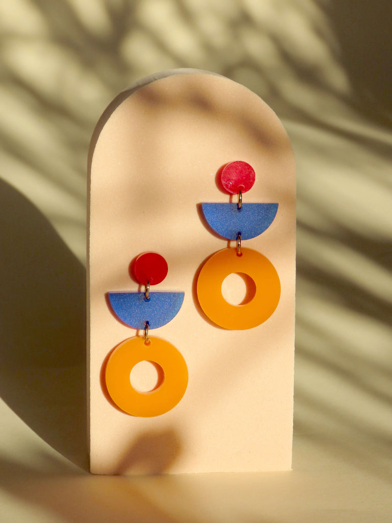 Verspielte Hängeohrringe aus Acryl mit roten runden Steckern, blauen Halbkreisen und großen orangefarbenen Ringen als Highlight