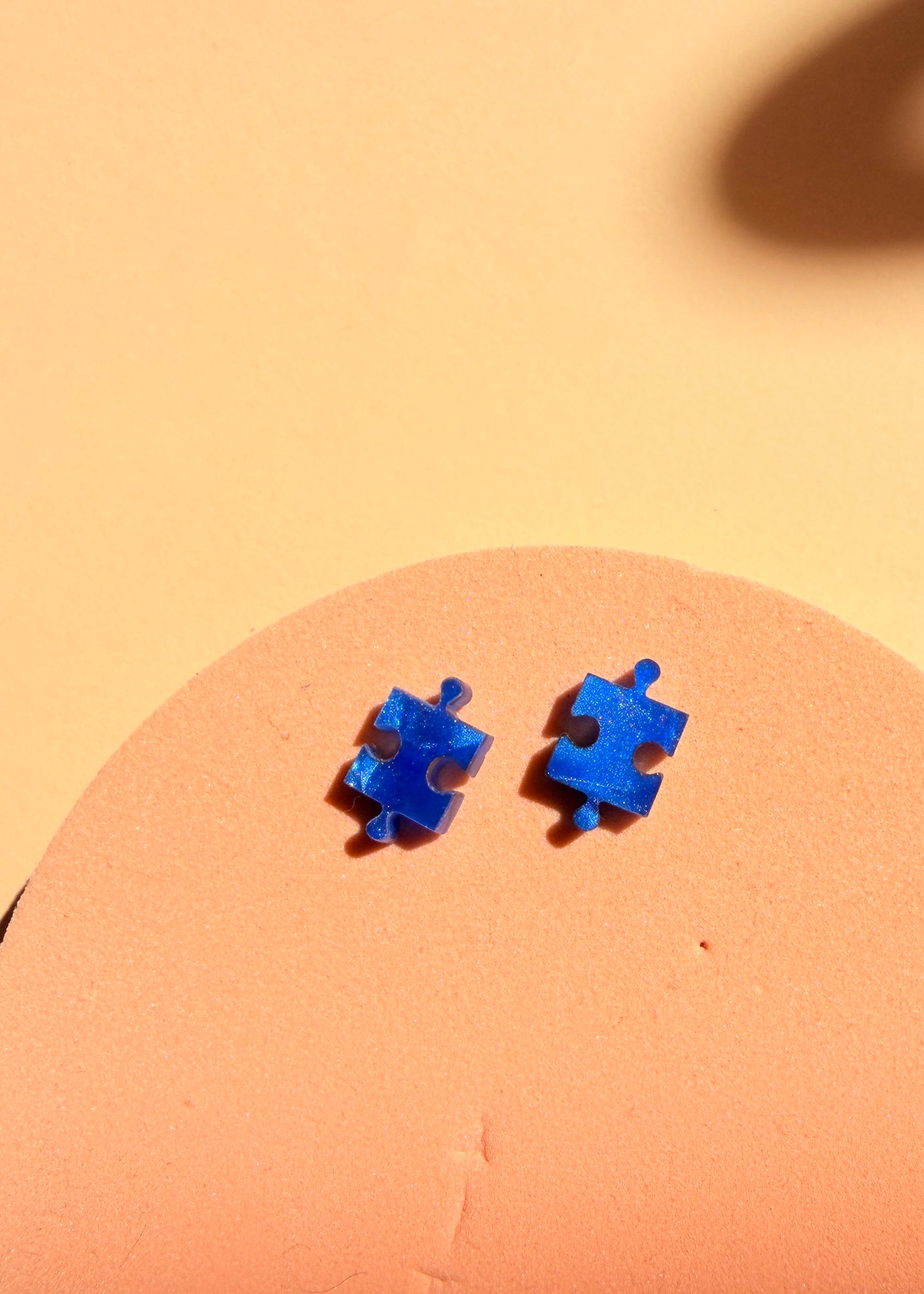 Ozeanblaue Puzzle Ohrstecker aus Edelstahl und Acryl – Detailansicht des Paares auf warmem Hintergrund
