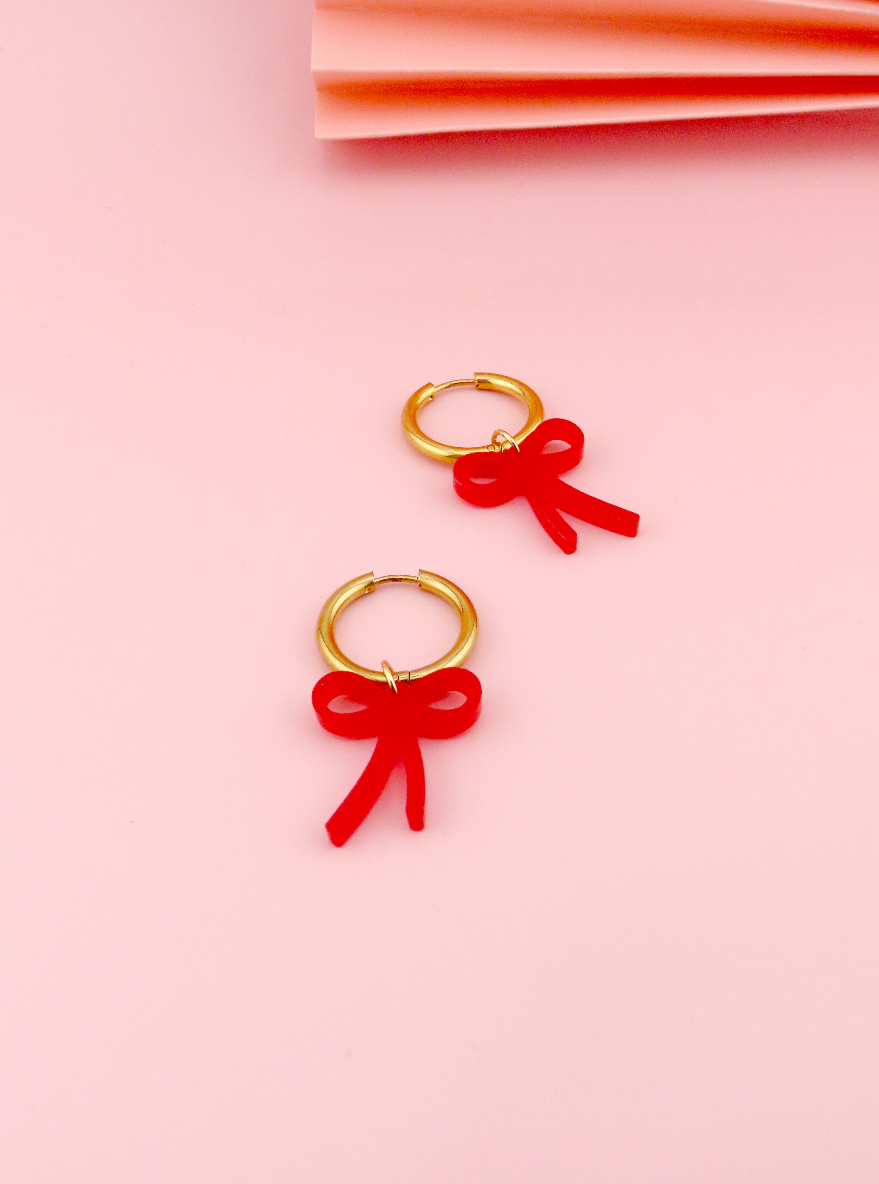 Vergoldete Creolen mit rotem Acrylanhänger in Schleifenform, leichtes Material und klare, verspielte Silhouette